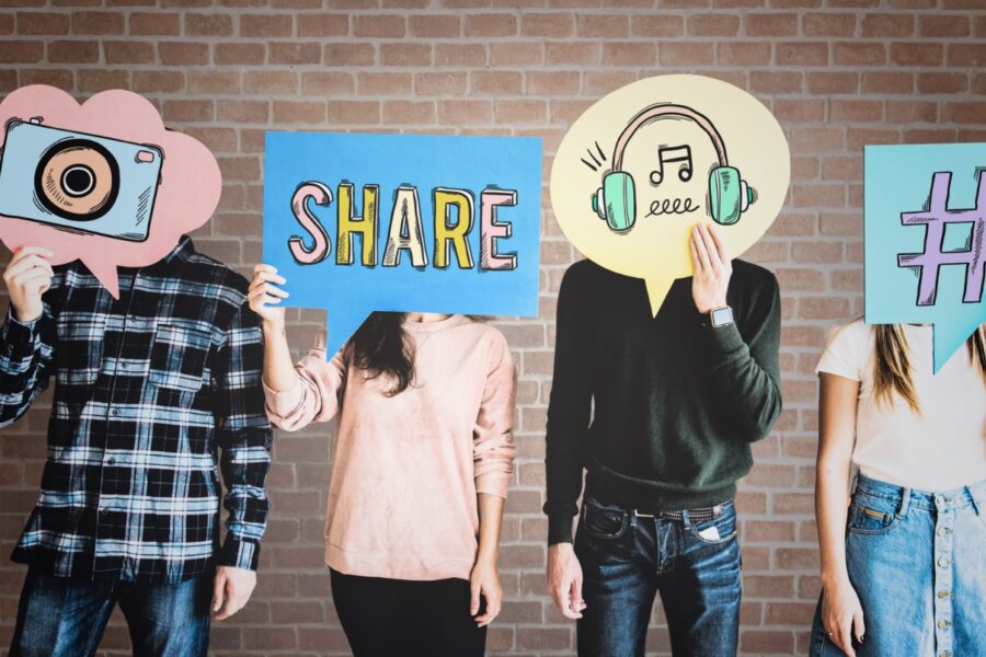 This picture shows four people standing side by side in front of a brick wall. Each person is holding a large, colorful sign in front of their face, so their faces are hidden. The signs are shaped like speech bubbles and contain symbols or words that represent social media and communication.